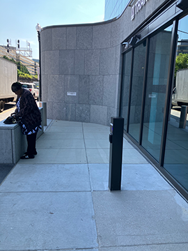 A picture of a building and some sidewalk. There is an automatic door opener slightly to the right of center in the foreground. To the left there are some windows, while to the right there is a woman at a half wall. In the center of the background there is a curved wall of the building.